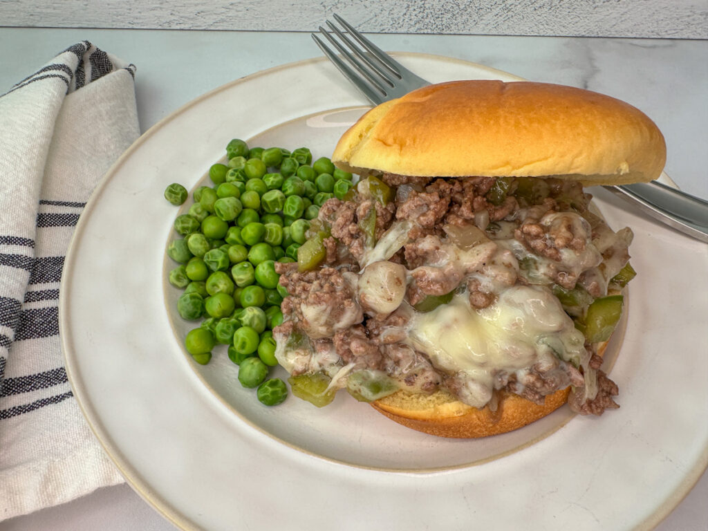 Sloppy joe sandwich on a bun with ground beef, green peppers, onion, and provolone cheese with peas on the side of the plate