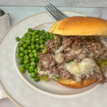 Sloppy joe sandwich on a bun with ground beef, green peppers, onion, and provolone cheese with peas on the side of the plate