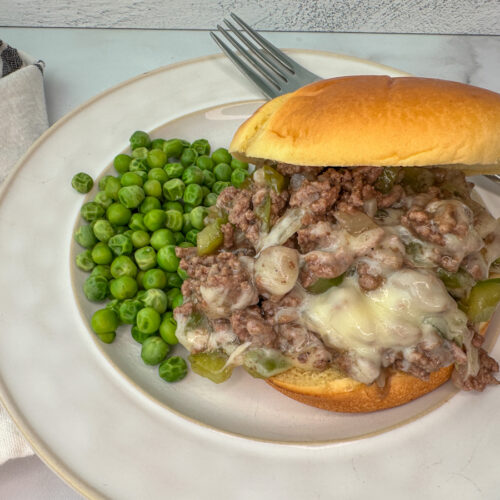 Sloppy joe sandwich on a bun with ground beef, green peppers, onion, and provolone cheese with peas on the side of the plate