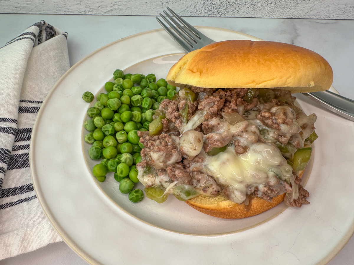 Sloppy joe sandwich on a bun with ground beef, green peppers, onion, and provolone cheese with peas on the side of the plate