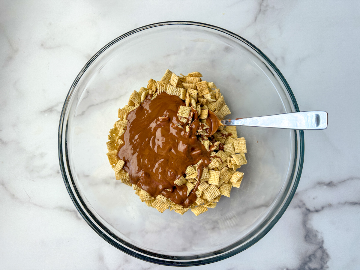 Chex mix in a large clear bowl with chocolate peanut butter liquid mixture on top