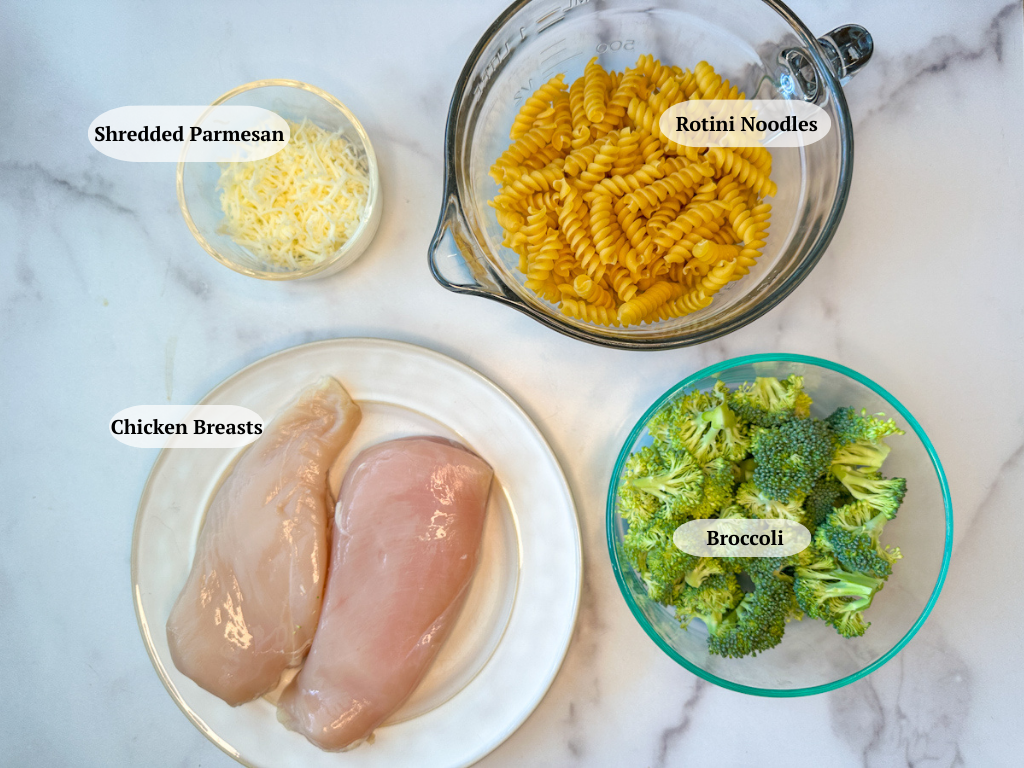 Chicken Alfredo with broccoli ingredients: A plate with two large chicken breasts, a bowl of broccoli florets, a bowl of rotini noodles, and a bowl of shredded parmesan cheese.