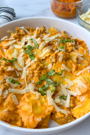 Creamy pumpkin ravioli with sausage on a plate topped with shredded parmesan cheese and basil.