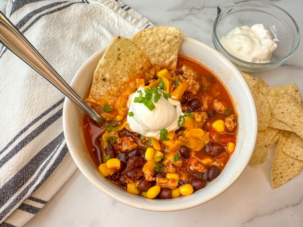 White bowl filled with easy turkey taco soup with a dollop of sour cream and sliced green onions on top with 2 tortilla chips as garnish.