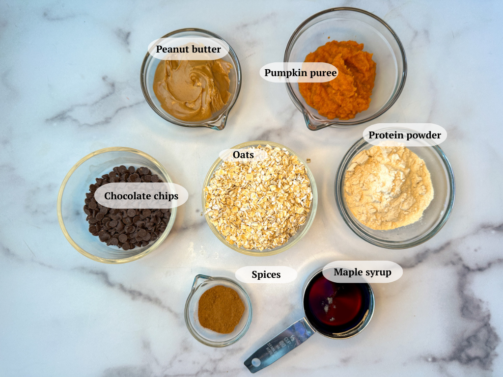 Pumpkin protein balls ingredients: pumpkin puree, vanilla protein powder, a bowl of peanut butter, a bowl of oats, a bowl of mini chocolate chips, a bowl of spices, and a measuring cup of maple syrup