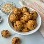 12 Protein Pumpkin Balls in a white bowl with a small bowl of mini chocolate chips and a bowl of oats.