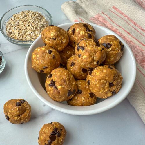 12 Protein Pumpkin Balls in a white bowl with a small bowl of mini chocolate chips and a bowl of oats.
