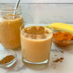 Pumpkin protein smoothie in a 12 oz glass with a mason jar of another smoothie, a bowl of pumpkin puree, a banana, and pumpkin pie spice in a small dish