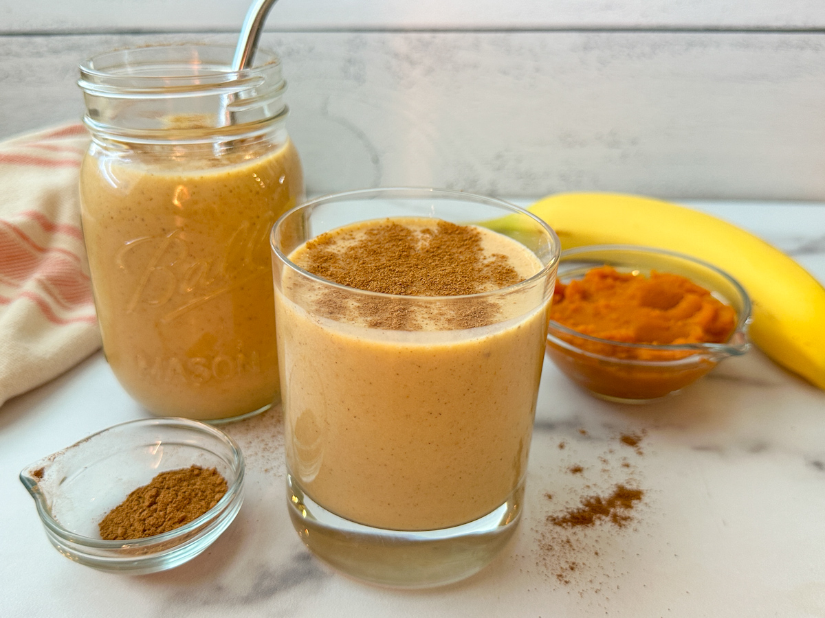 Pumpkin protein smoothie in a 12 oz glass with a second smoothie in a mason jar. There's a small jar of pumpkin pie spice with a small bowl of pumpkin puree beside a banana. 