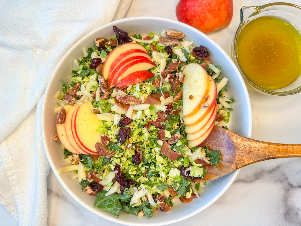 a large white bowl filled with a fall brussels sprouts and apple salad with shredded brussels, chopped kale, sliced apples, and dried cranberries.