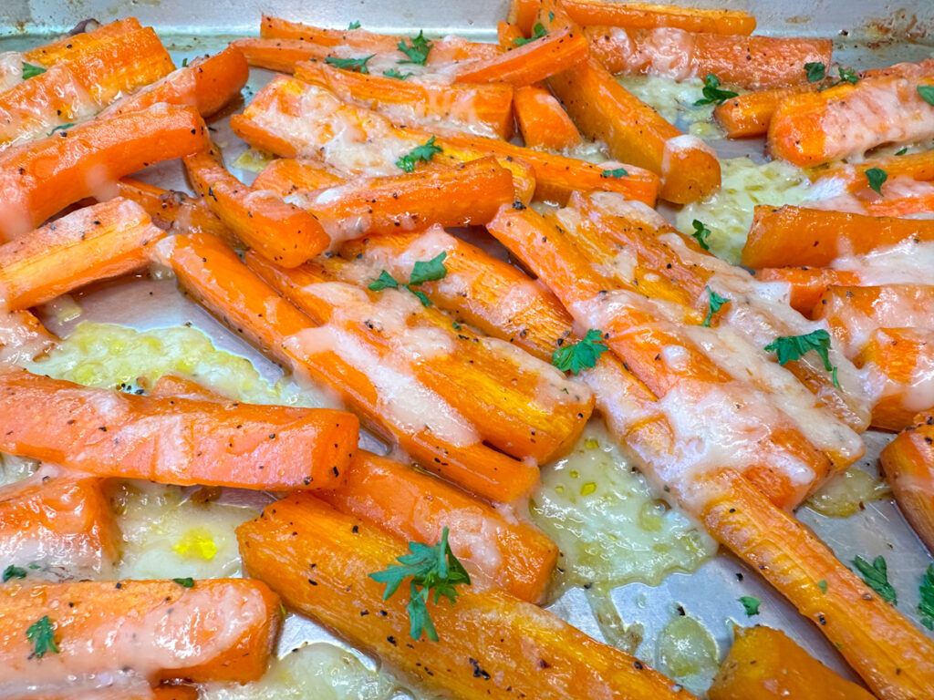 Quartered carrot sticks with melted parmesan cheese topped with chopped parsley on a baking sheet.
