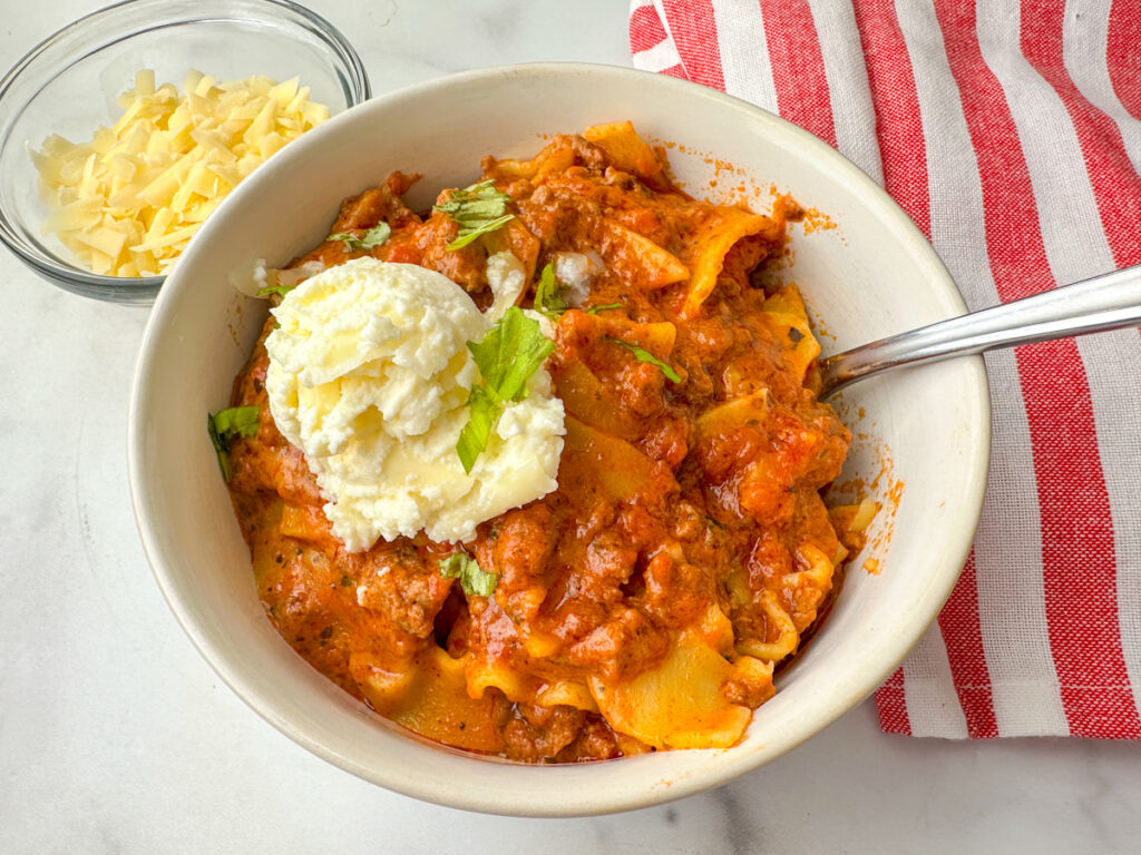 bowl of lasagna soup with cooked lasagna noodle pieces, cooked ground beef, tomato sauce, and a dollop of ricotta cheese on top.