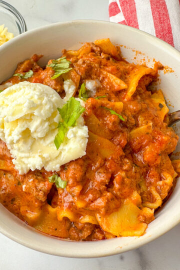 bowl of lasagna soup with cooked lasagna noodle pieces, cooked ground beef, tomato sauce, and a dollop of ricotta cheese on top.