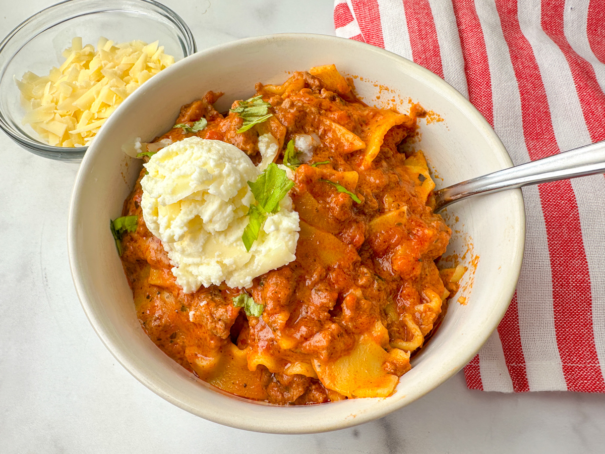 Bowl of slow cooker lasagna soup with a dollop of ricotta cheese on top and a small bowl of shredded mozzarella beside it.