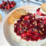 whipped cranberry feta dip on a large plate topped with chopped pecans. Someone is dipping a cracker in the dip.