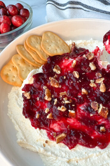 whipped cranberry feta dip on a large plate topped with chopped pecans. Someone is dipping a cracker in the dip.
