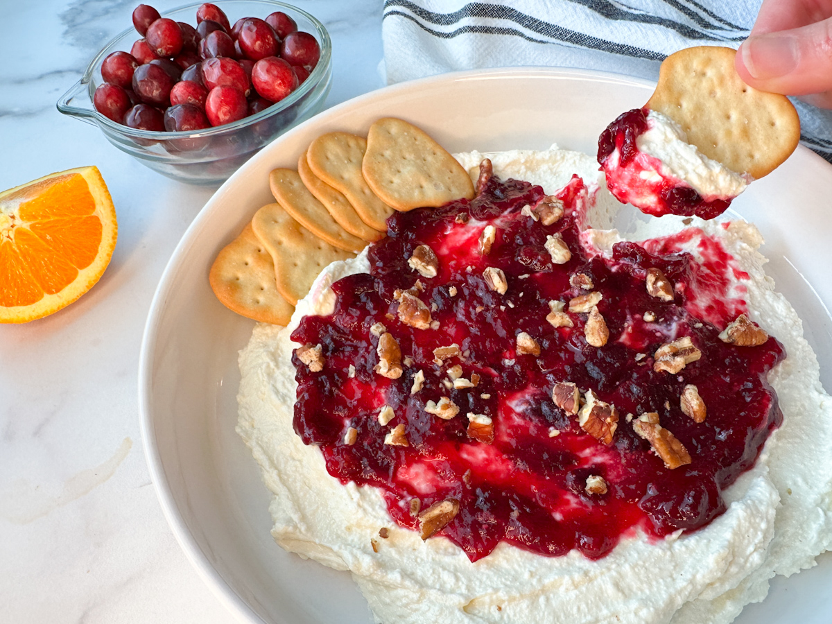 Cranberry whipped feta dip on a large plate topped with smooth cranberry sauce and chopped pecans. Crackers on the side for dipping.