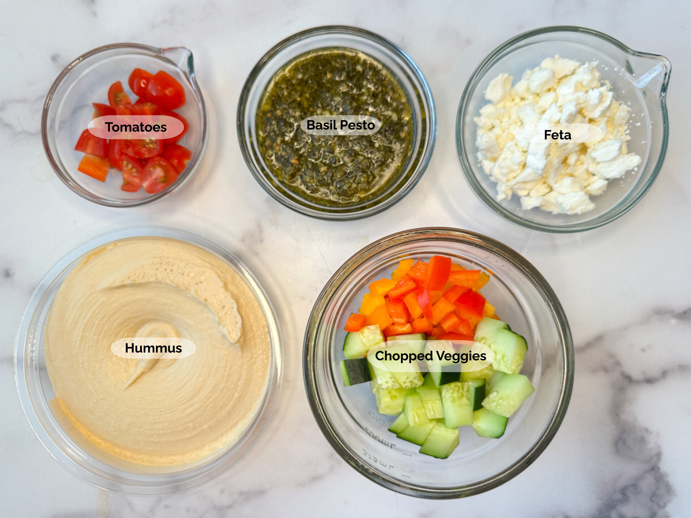 Greek Hummus dip ingredients: plain hummus, chopped cherry tomatoes, basil pesto, feta, and chopped cucumbers, red bell and orange bell peppers.