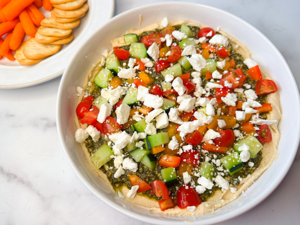 Easy Greek Hummus Dip on a white serving platter. Hummus topped with pesto topped with chopped veggies and feta cheese.