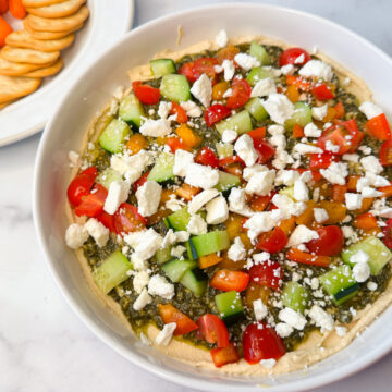 Easy Greek Hummus Dip on a white serving platter. Hummus topped with pesto topped with chopped veggies and feta cheese.