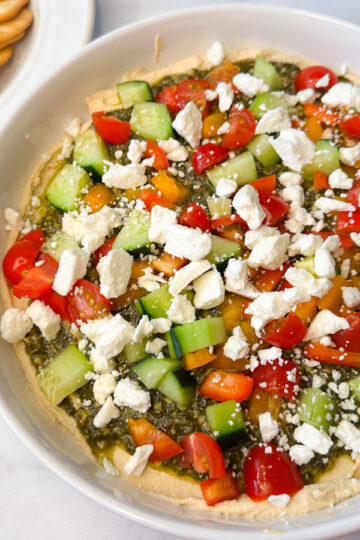 Easy Greek Hummus Dip on a white serving platter. Hummus topped with pesto topped with chopped veggies and feta cheese.