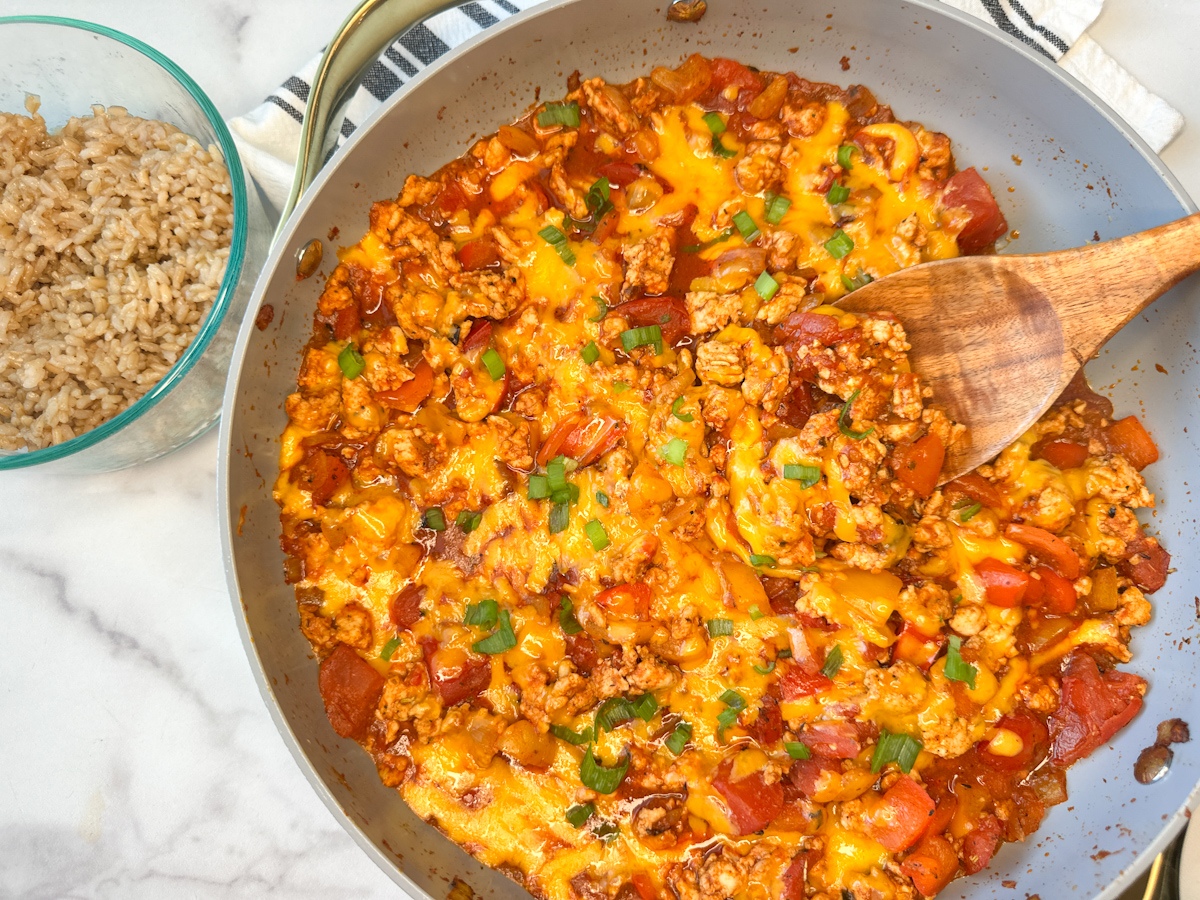 Cooked unstuffed pepper skillet meal in a large skillet with a side of rice. Cooked turkey with chopped cooked peppers, shredded, melted cheese, and diced tomatoes topped with sliced green onions.