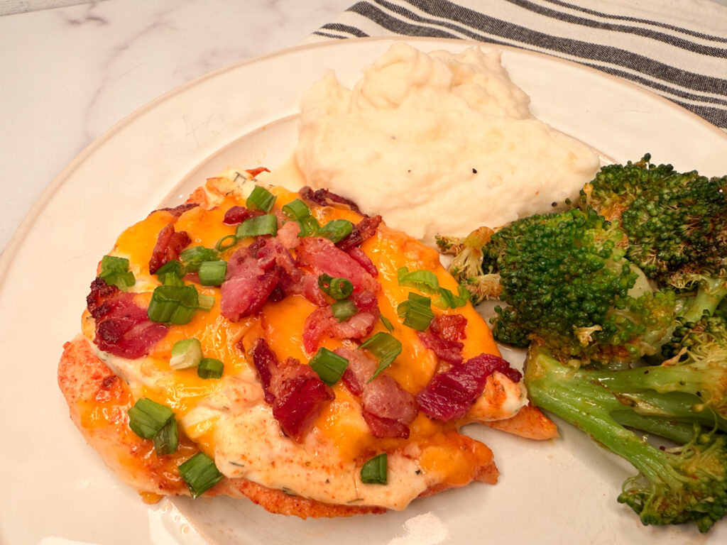 Baked Crack Chicken topped with shredded cheese, crumbled bacon, and green onion slices with a side of broccoli and mashed potatoes.