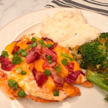 Baked Crack Chicken topped with shredded cheese, crumbled bacon, and green onion slices with a side of broccoli and mashed potatoes.