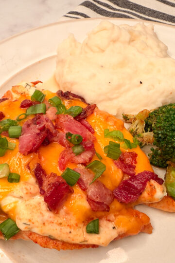 Baked Crack Chicken topped with shredded cheese, crumbled bacon, and green onion slices with a side of broccoli and mashed potatoes.