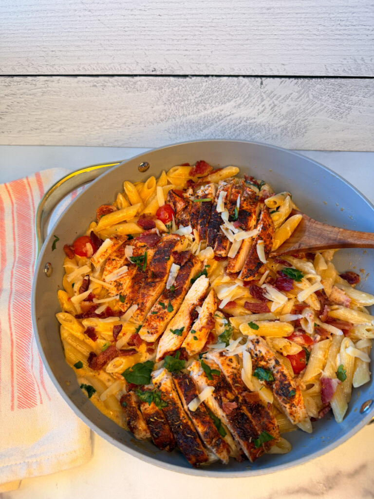 Creamy bacon and chicken pasta cooked in a large skillet with cooked noodles in a cheesy sauce topped with grilled chicken and crumbled bacon.