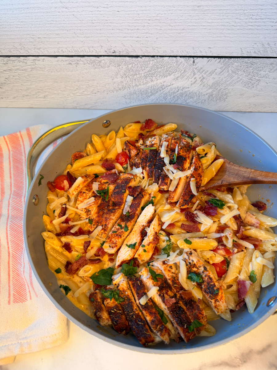 Creamy Bacon & Spinach Pasta in a large skillet. Cooked penne noodles topped with a creamy sauce, sliced grilled chicken, and bits of bacon and diced tomato. 