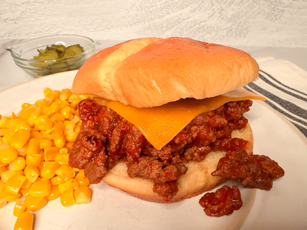 Sloppy Joe sandwich with saucy meat spilling out of bun. Meat is topped with a slice of cheese and there's a side of corn on the side.