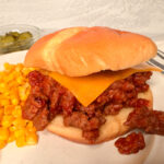 Sloppy Joe sandwich with saucy meat spilling out of bun. Meat is topped with a slice of cheese and there's a side of corn on the side.