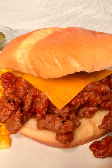 Sloppy Joe sandwich with saucy meat spilling out of bun. Meat is topped with a slice of cheese and there's a side of corn on the side.