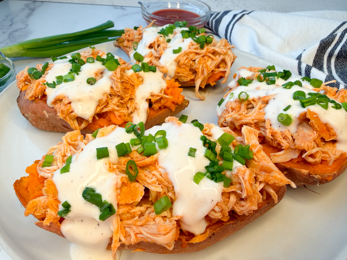 4 sweet potatoes on a plate topped with a shredded buffalo chicken mixture and topped with ranch and sliced green onions.