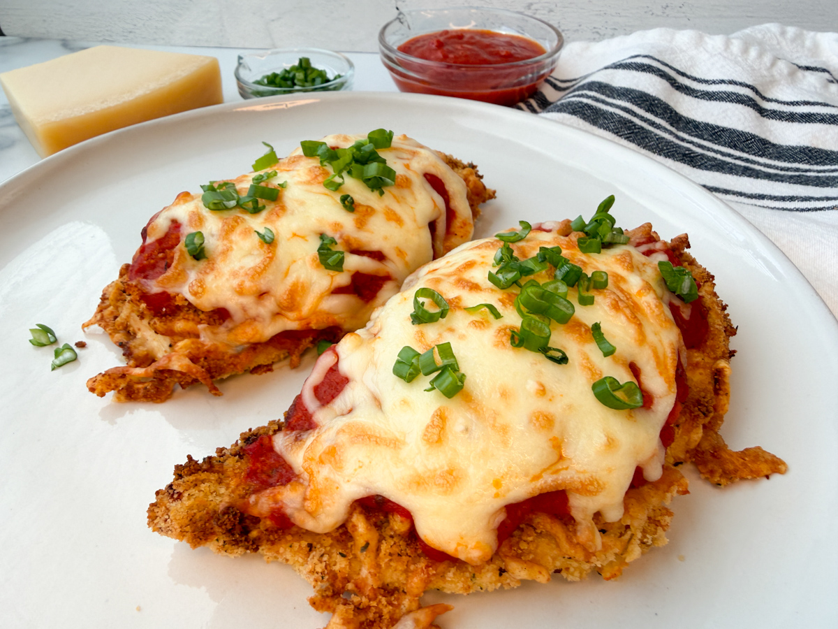 Air Fryer Chicken Parmesan: 2 crispy breaded chicken breasts coated in breadcrumbs and topped with marinara and melted mozzarella cheese. Topped with sliced green onions.