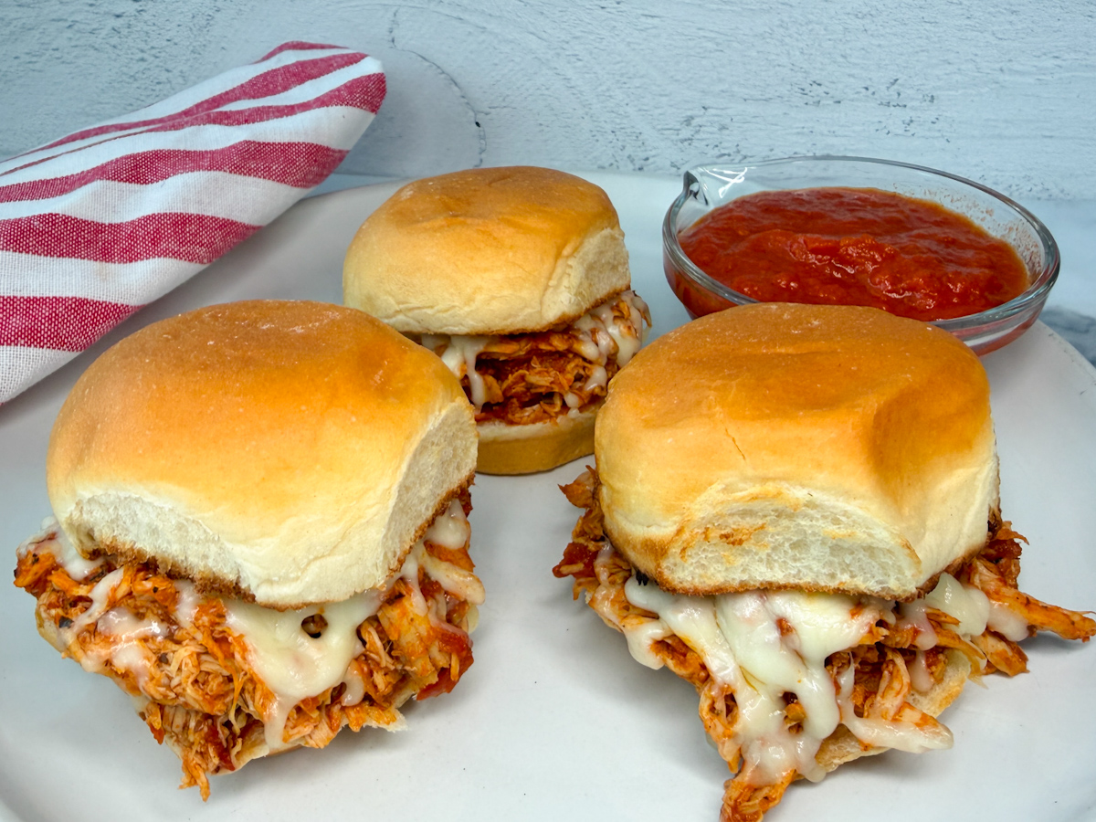 3 mini chicken parmesan sliders on a plate with a side of marinara sauce. Sandwiches are filled with saucy, shredded chicken with melted cheese.