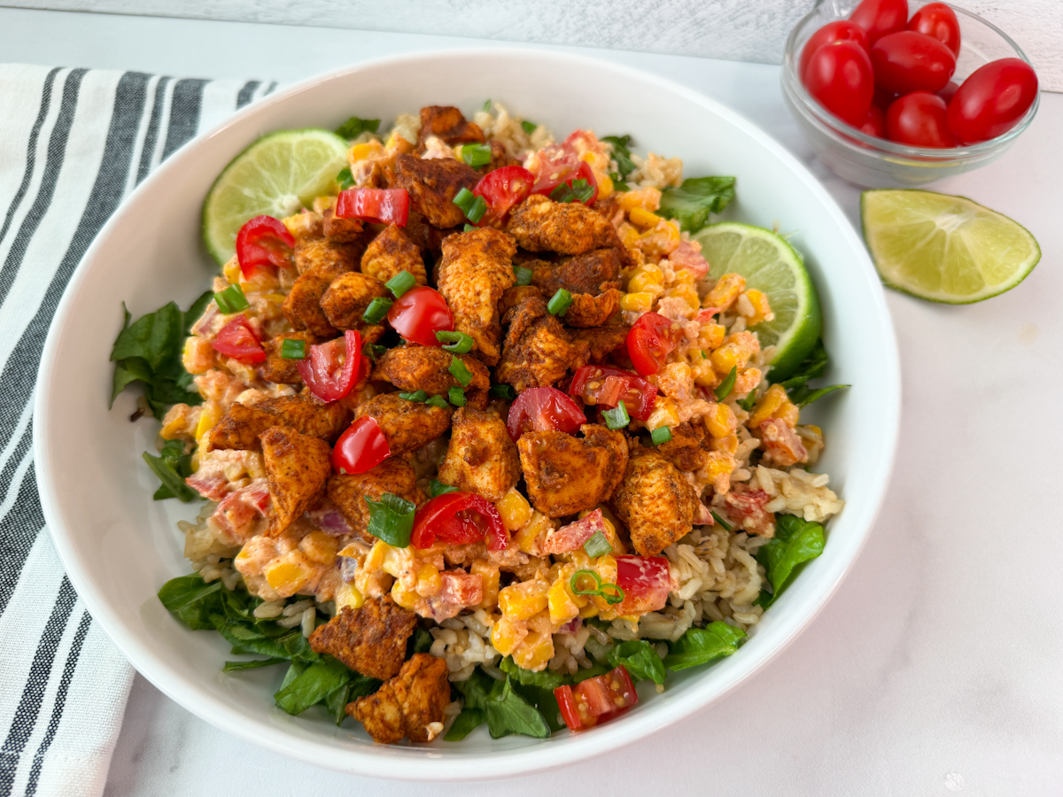 Street Corn Chicken Rice Bowl in a large bowl with romaine, corn, grilled chicken, tomatoes, rice, and lime wedges.