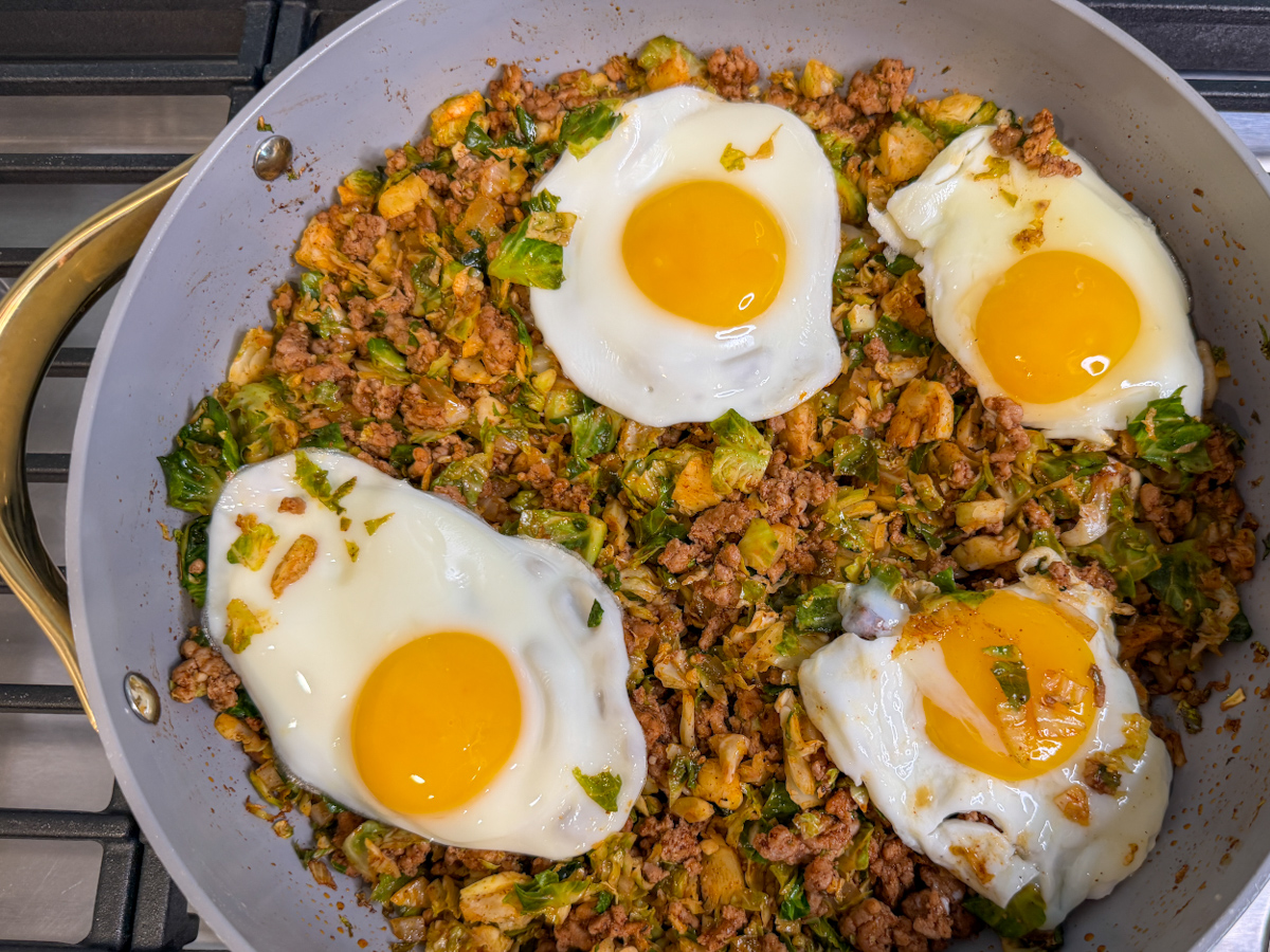 Spicy Pork Brussels Bowl in a large skillet with cooked pork, shredded brussels, and topped with 4 over easy eggs.